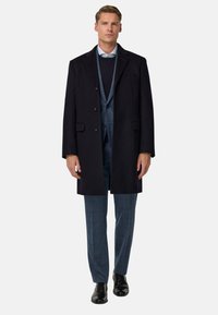 Námornícky vlnenný kabát s veľkými prednými gombíkmi, skombinovaný s tmavým vestom a svetlomodrou košeľou. Model má na sebe textúrované svetlomodré nohavice a čierne topánky.