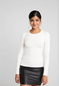 Haut blanc à manches longues côtelé avec un col rond, assorti à une mini-jupe en cuir noir ajustée. Texture lisse, design ajusté.