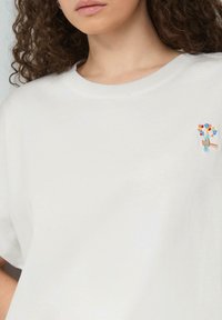 T-shirt en coton blanc avec un design brodé sur la poitrine gauche représentant une main tenant un bouquet de fleurs colorées. Encolure ronde et manches courtes.