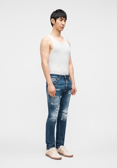 Λευκό αμάνικο τοπ, φθαρμένο μπλε τζιν με διάφορες σκισίματα, ανοιχτόχρωμα παπούτσια με διακριτικές λεπτομέρειες· μοντέλο που στέκεται πλευρικά σε ουδέτερο φόντο.