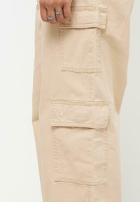 Beige cargobyxor med flera sidofickor, sömnadsdetaljer och en broderad logotyp i vitt på den övre fickan. Mjuk bomullsmaterial.