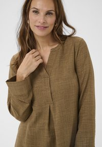 Túnica marrón con textura, escote en V y mangas largas. Presenta un pliegue frontal y un ajuste relajado. La tela parece ligera y transpirable.