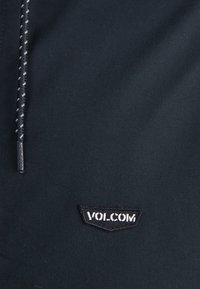 Tela negra con una textura suave, que presenta un parche con el logo bordado en blanco y negro que dice "VOLCOM." Cordon delgado trenzado.