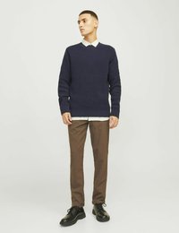 Marinblå stickad tröja med V-ringning, krämfärgad skjorta under, bruna tapered byxor och svarta snörskor, stående mot en neutral bakgrund.