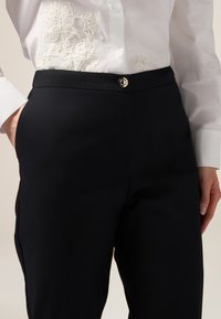 Černé tailored kalhoty s hladkou texturou, s přední kapsou, zapínáním na zip a kulatým kovovým knoflíkem jako akcentem.