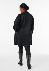 Schwarzer Trenchcoat mit lockerer Passform, langen Ärmeln und seitlichen Taschen. Über schwarzen Leggings und kniehohen schwarzen Stiefeln getragen. Glatte Textur.