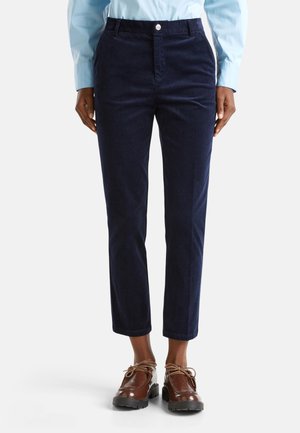 Persoon in donkerblauwe corduroy broek, lichtblauw shirt en bruine veterschoenen, staand tegen een effen witte achtergrond.