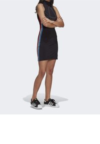 Černé bezrukávové šaty s vysokým límcem, s červenými a modrými postranními pruhy. Spárováno s černobílými teniskami. Hladká textura.