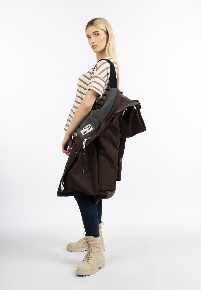 Cappotto marrone con cerniere e colletto imbottito grigio, indossato su una maglietta a righe. Abbinato a jeans scuri e stivaletti chiari con lacci.