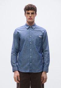 Modra chambray srajca z dolgimi rokavi, ovratnikom na gumbe, prsnim žepom in logotipnim obližem. Gladek tekstura in prilegajoč kroj.