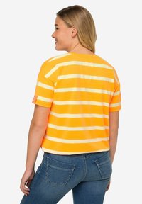 Camiseta de manga corta a rayas en amarillo brillante y blanco, con un corte relajado y una etiqueta en la manga izquierda. Combinada con jeans de mezclilla azules.