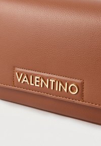 Smeđa kožna torbica s teksturom nalik na šljunak i zlatnim logom "VALENTINO". Ima elegantan pravokutni dizajn s detaljima ušivenim u tkaninu.