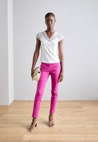 Haut blanc avec dentelle, jeans roses et escarpins blancs à lanières. Le mannequin tient un sac à main crème avec des accents en chaîne. Fond en parquet.