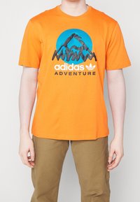 Oranžové bavlněné tričko s modrým kruhovým grafikem s horskými vrcholy a černým nápisem "adidas ADVENTURE". Standardní střih, kulatý výstřih.