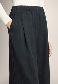 Črne hlače z drobnimi črtami in elastičnim pasom, z bočnimi žepi in ohlapnim krojem. Tkanina je videti gladka in strukturirana.