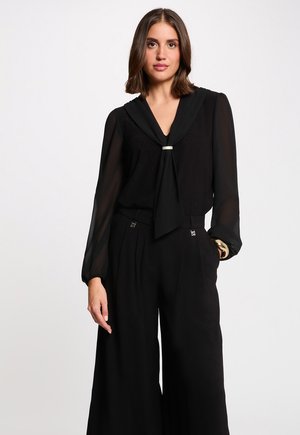 Vrouw met schouderlang bruin haar, gekleed in een zwarte doorschijnende blouse met lange mouwen en een stropdas, en wijde zwarte broek tegen een witte achtergrond.