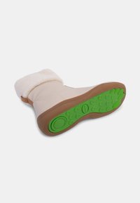 Crème-kleurige laars met een zachte, harige rand, tan-kleurige zijkanten en een bruine rubberen zool met een felgroene, gestructureerde onderkant.