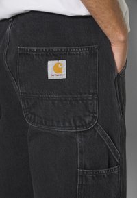Schwarze Denim-Hose mit einer geräumigen Gesäßtasche, die ein weißes und gelbes Carhartt-Logo trägt. Der Stoff hat ein leicht strukturiertes Aussehen.