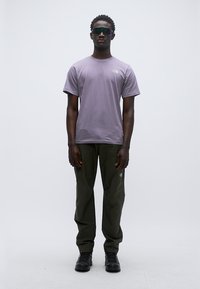 Μπλούζα με κοντά μανίκια σε λιλά χρώμα και παντελόνι σκούρο πράσινο. Το μοντέλο φοράει σκούρα γυαλιά ηλίου και μαύρα παπούτσια. Η μπλούζα έχει ένα λογότυπο στο στήθος.