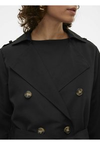Black trench coat with a double-breasted design, large lapels, and glossy buttons. Smooth fabric texture, seen partially with a black top beneath.