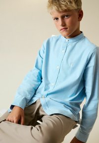 Hellblaues Langarmhemd mit Stehkragen, vorne mit Knöpfen und einem kleinen gestickten Logo auf der Brust.