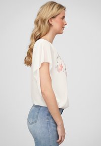 Camicia bianca con maniche a sbuffo corte, stampa floreale in rosa e grigio, abbinata a jeans azzurri chiari. Tessuto liscio e leggero.