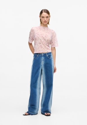 Nainen pukeutuneena vaaleanpunaiseen kukalliseen lyhythihaiseen toppiin, korkeavyötäröisiin leveälahkeisiin sinisiin farkkuihin ja mustiin avokärkisiin sandaaleihin, seisomassa valkoista taustaa vasten.