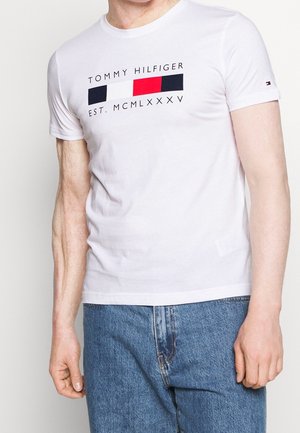 Biała koszulka Tommy Hilfiger z logo w kolorach czerwonym, białym i niebieskim oraz napisem "EST. MCMLXXXV", noszona przez osobę w niebieskich dżinsach.