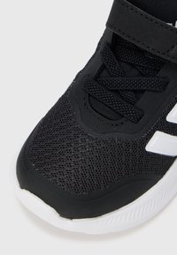 Črni športni čevelj z zgornjim delom iz mrežastega materiala, teksturiranimi materiali, gumijastim podplatom in belimi poudarki. Trak z ježkom omogoča enostavno uporabo.