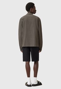 Olive green, lightweight jacket with long sleeves, straight cut, and back yoke detail, paired with black shorts and black sandals.
