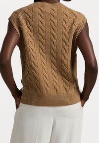 Neuleliivi vaaleanruskeana, jossa on resorireuna ja palmikkoneulekuvio; hihaton malli paljastaa hartiat ja kädet.