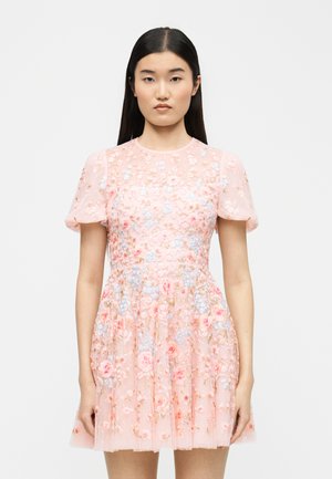 Młoda kobieta z długimi ciemnymi włosami, ubrana w krótką różową sukienkę z bufkami na rękawach i wielokolorowym haftem kwiatowym, stojąca na jednolitym tle.