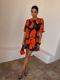 Un vestito floreale con papaveri arancioni su sfondo nero, maniche a sbuffo corte, texture plissettata e gonne a strati, abbinato a una borsa nera.