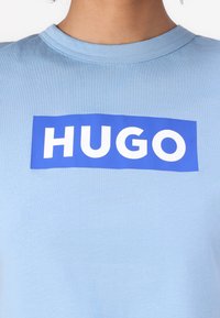 Šviesiai mėlynos spalvos medvilninė marškinėliai su ryškiu mėlynu stačiakampiu logotipu, kurioje baltu šriftu yra žodis "HUGO", esantis centre priekyje. Trumpomis rankovėmis ir apvaliu iškirpimu.