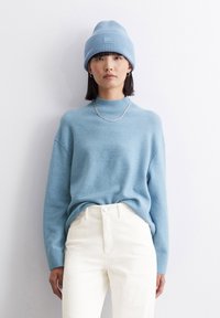 Pulover tricotat de culoare bleu deschis cu guler înalt, asortat cu pantaloni de corduroy crem. Modelul poartă o căciulă albastră asortată și un colier din lanț argintiu.