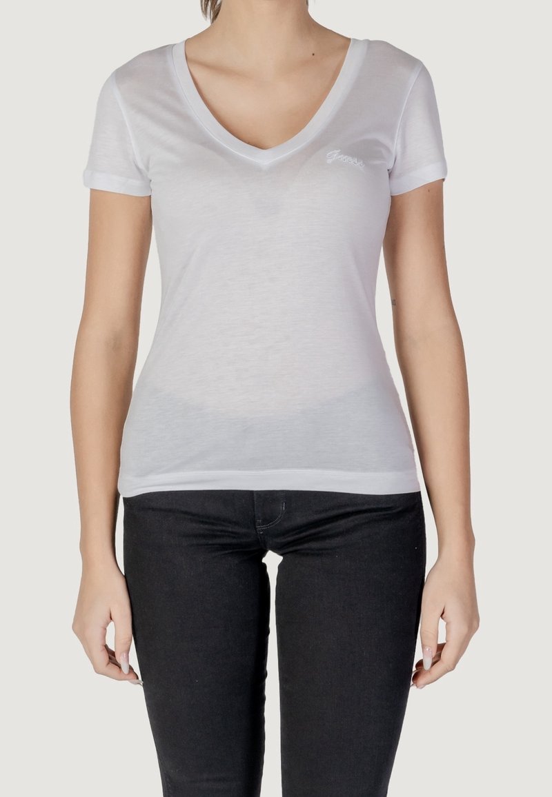 T-shirt blanc à col en V, manches courtes, tissu léger et légèrement transparent, avec une broderie subtile sur la poitrine, associé à un jean foncé.