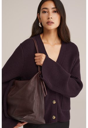 Giovane donna che indossa un cardigan a maglia viola scuro e orecchini a cerchio dorati, mentre tiene una grande borsa di pelle marrone scuro appesa alla spalla.