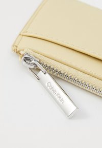 Portefeuille en simili cuir beige avec une tirette en métal argenté gravée "Calvin Klein", texture lisse et design minimaliste.