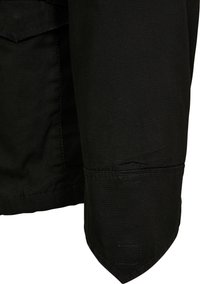 Zwarte stoffen jas-sleeve met een diagonale snit, details van bovenstiksels en een klein rechthoekig patch dicht bij de manchetten.