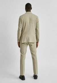 Ljus beige kostym med strukturerad tyg, skräddarsydd passform och två bakventiler. Matchad med svarta skor, sedd bakifrån.