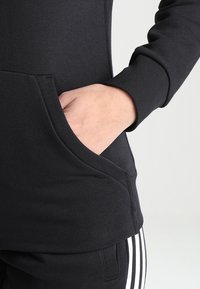 Sweatshirt noir avec une poche kangourou, des poignets côtelés et une texture lisse, associé à un pantalon noir avec des rayures blanches sur les côtés.