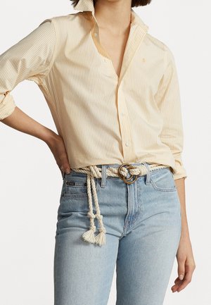 Frau trägt ein hellgelb gestreiftes Hemd mit hochgekrempelten Ärmeln, hellblaue Jeans und einen weißen Seilgürtel mit goldener Schnalle.