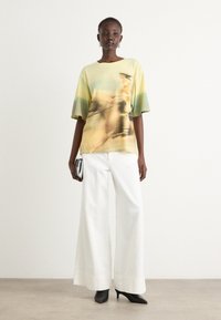 Loose-fitting short-sleeve shirt in yellow and green hues, featuring an abstract print. Paired with wide white trousers and black heels.