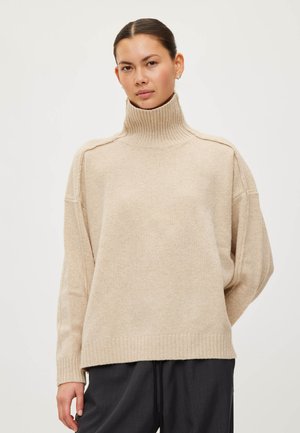 Femme aux cheveux foncés portant un pull à col roulé côtelé beige et un pantalon foncé, debout devant un fond clair uni.