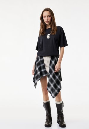 Junge Frau trägt ein schwarzes, übergroßes T-Shirt mit einem Hundemotiv, einen schwarz-weiß karierten Rock und schwarze Stiefel mit Socken bis zur Mitte der Wade.