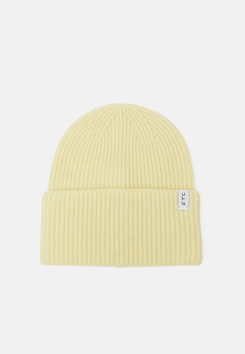Gul ribbestrikket lue med en brettet kant, med et lite hvitt stoffmerke med svart tekst. Myk tekstur og tettsittende design.