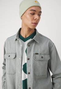 Camisa de mezclilla gris con dos bolsillos en el pecho, superpuesta a un suéter blanco y verde con estampado, rematada con un gorro ligero verde que lleva un logo.
