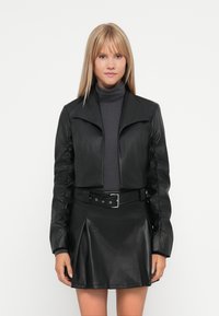 Svart faux skinn cropped jakke med bred krage og rynkete ermer, sammen med en matchende plisserte skjørt og grå turtleneck.