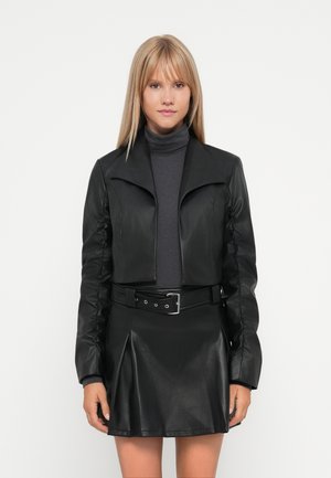 Black faux leather cropped jacket with a wide collar and gathered sleeves, paired with a matching pleated skirt and gray turtleneck.