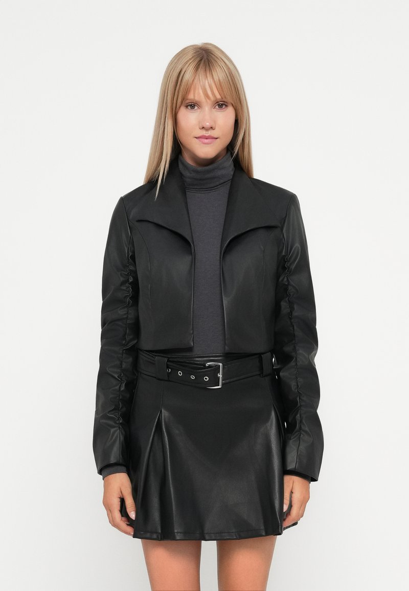 Svart faux skinn cropped jakke med bred krage og rynkete ermer, sammen med en matchende plisserte skjørt og grå turtleneck.
